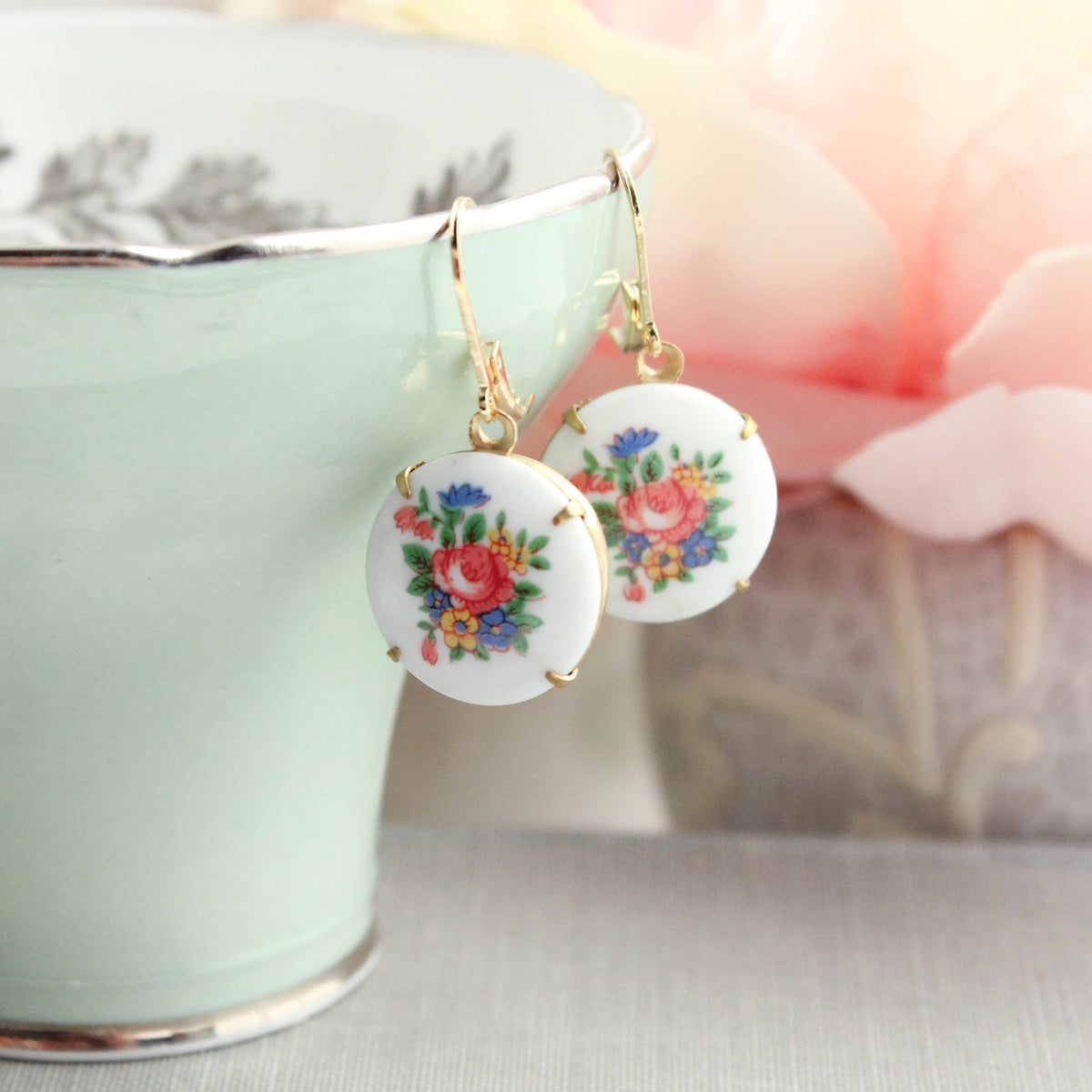 A pair of round white drop earrings with colorful floral designs and gold hooks, hanging from the rim of a pale green teacup with soft pink flowers in the background.