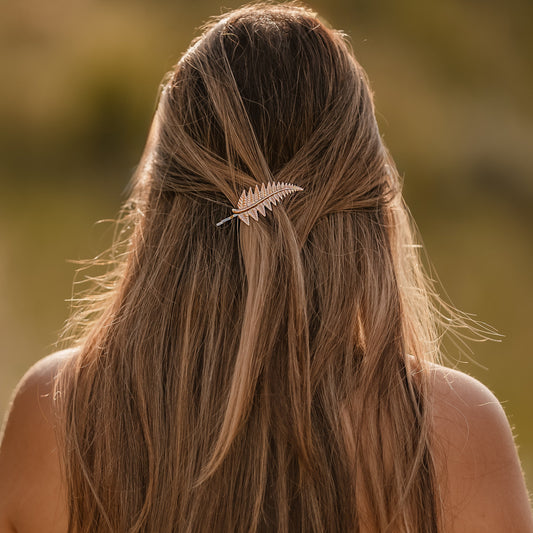 A woman viewed from behind with long, straight light brown hair, wearing a delicate gold feather hair clip. She is wearing an off-the-shoulder floral dress against a soft-focus outdoor background.