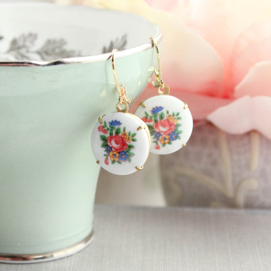A pair of round white drop earrings with colorful floral designs and gold hooks, hanging from the rim of a pale green teacup with soft pink flowers in the background.