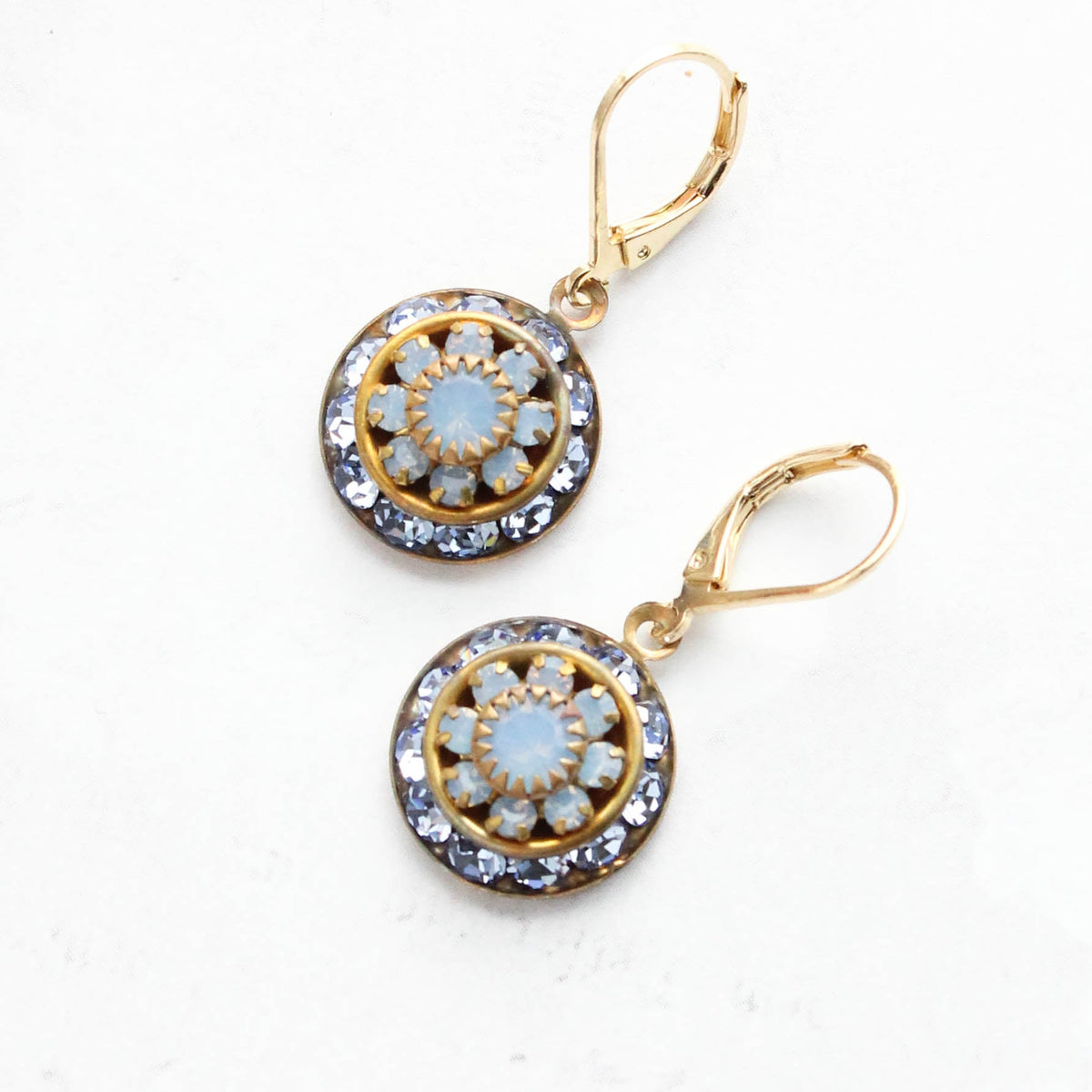 A pair of gold leverback drop earrings with circular floral designs featuring light blue opal-like center stones and clear rhinestone borders, displayed on a light textured background.