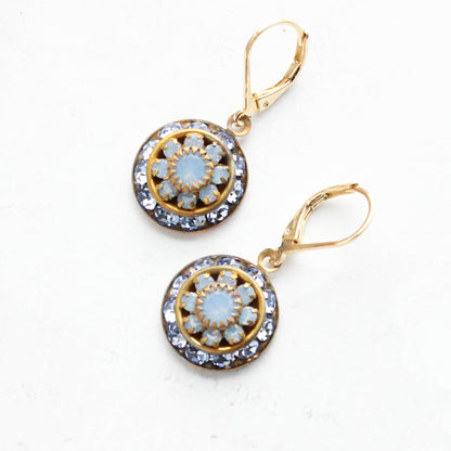 A pair of gold leverback drop earrings with circular floral designs featuring light blue opal-like center stones and clear rhinestone borders, displayed on a light textured background.