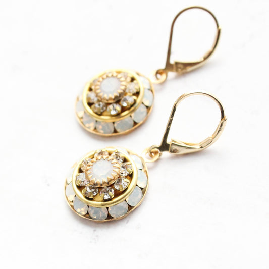 A pair of gold lever back earrings with circular, scalloped settings featuring a central milky white opal glass stone surrounded by small clear crystals and bordered by round white stones, shown on a plain white background
