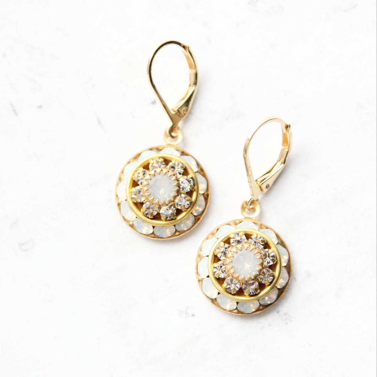 A pair of gold leverback drop earrings with circular floral designs, featuring white opal glass and clear rhinestone accents, displayed on a white background.