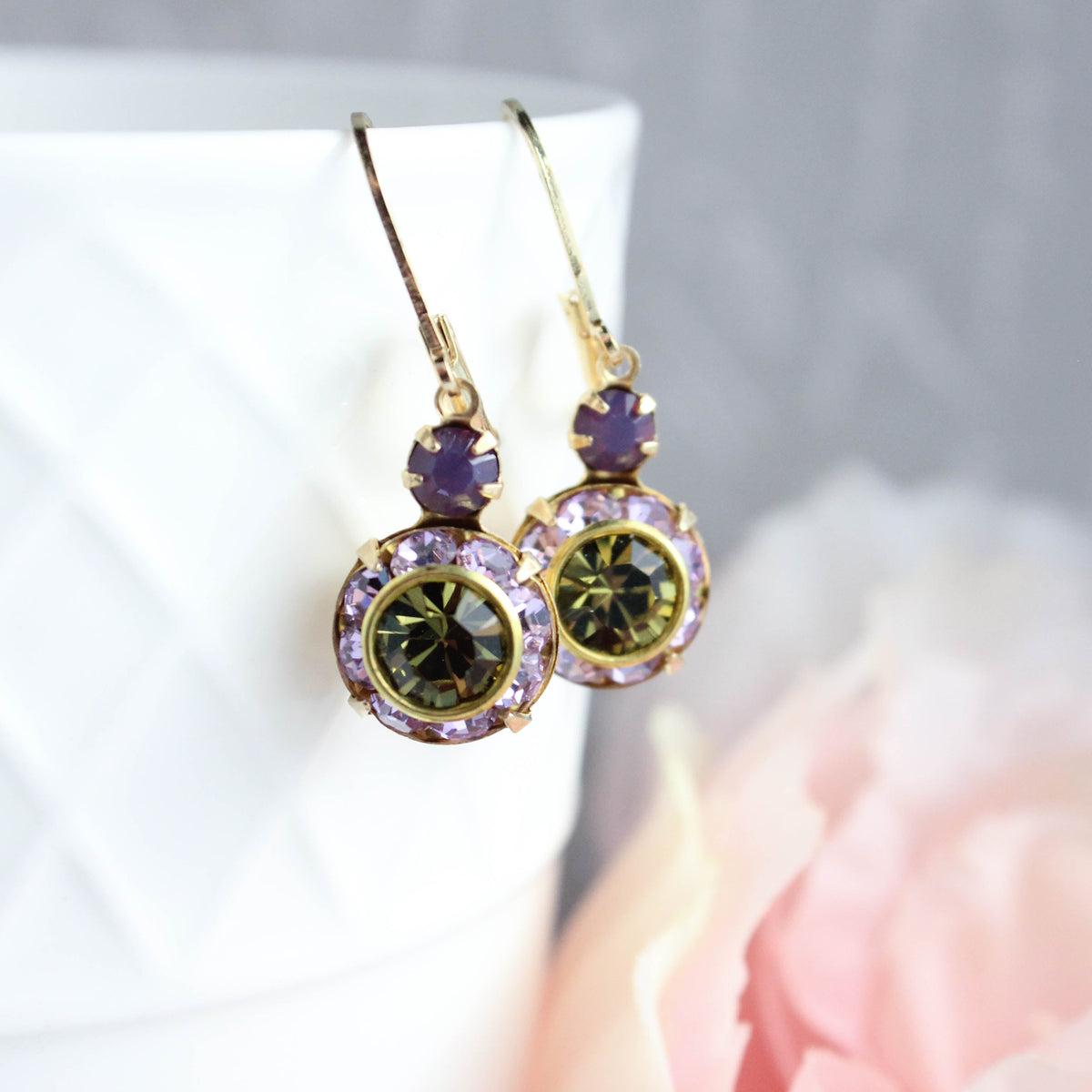 A pair of gold drop earrings with oval green gemstones surrounded by small lavender-purple crystals, displayed on a white textured surface with a soft pink flower in the foreground.