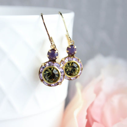 A pair of gold drop earrings with oval green gemstones surrounded by small lavender-purple crystals, displayed on a white textured surface with a soft pink flower in the foreground.