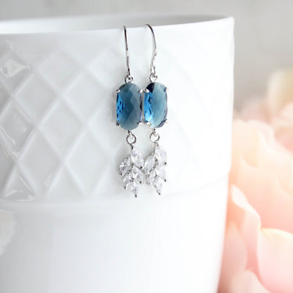 A pair of dangle earrings with blue faceted oval stones and silver-toned leaf-shaped crystal drops, hanging from silver hooks against a white textured ceramic cup with a soft pink blurred background.