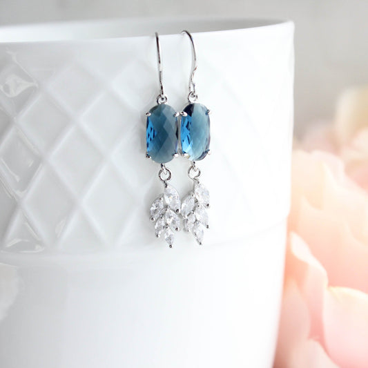 A pair of dangle earrings with blue faceted oval stones and silver-toned leaf-shaped crystal drops, hanging from silver hooks against a white textured ceramic cup with a soft pink blurred background.