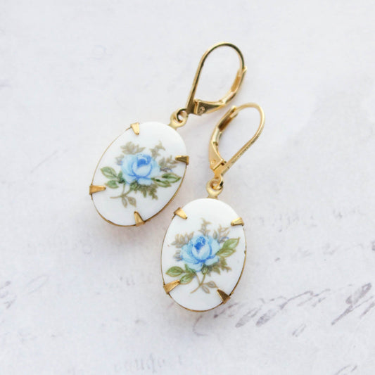A pair of gold leverback earrings with oval white enamel pendants featuring a delicate blue rose and green leaves, displayed on a light textured background.