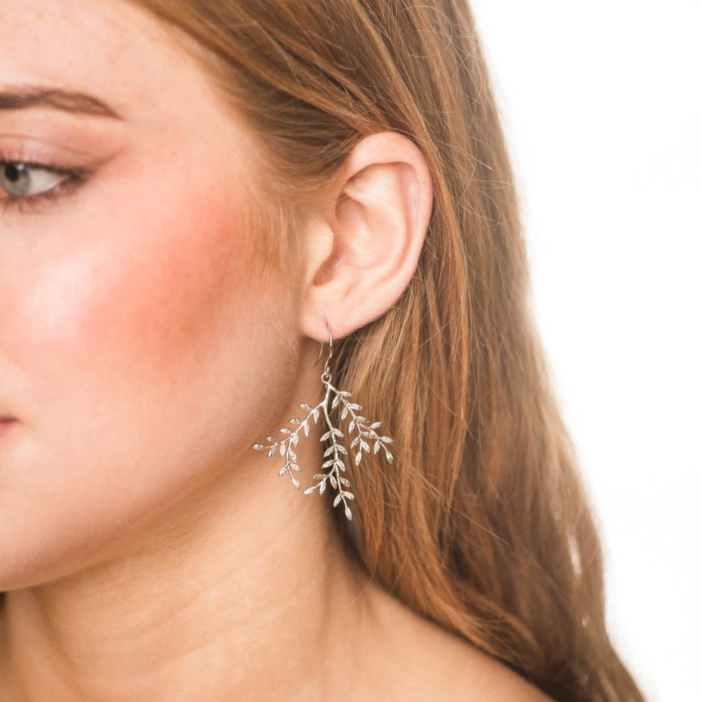 A close-up of a woman's ear and side profile wearing a delicate silver dangle earring shaped like small leaves and branches, against a plain white background.