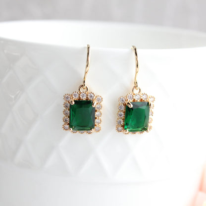 A pair of gold drop earrings with square emerald-cut green gemstones and small clear stones in a halo setting, displayed on a white textured ceramic cup.