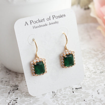 A pair of gold-tone drop earrings with square green gemstones and small clear stones, displayed on a white card labeled 'A Pocket of Posies Handmade Jewelry' on a white patterned surface.