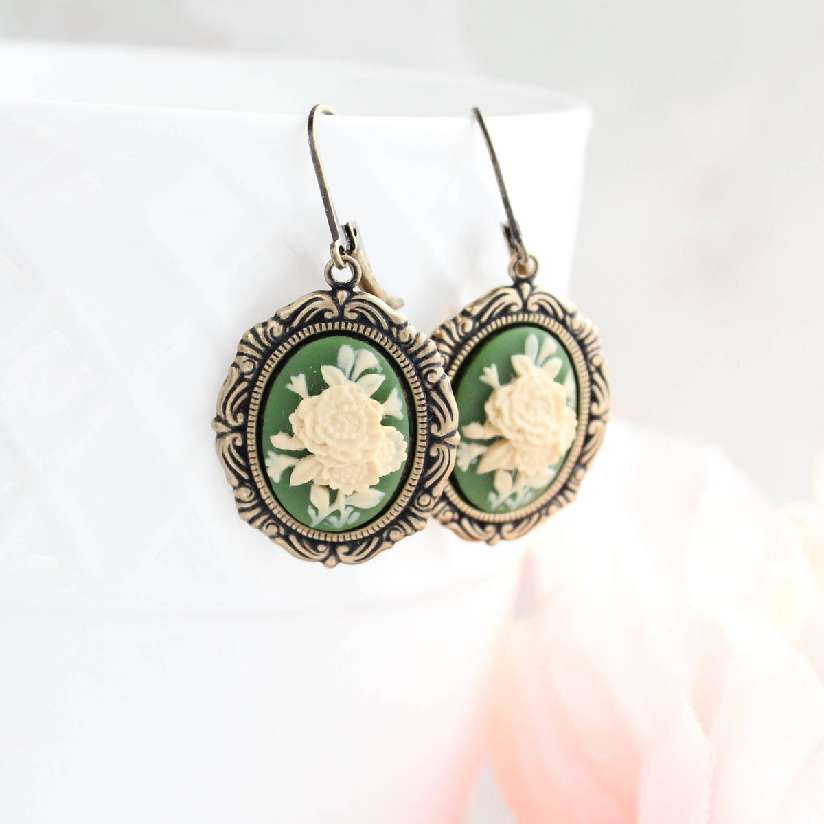 A pair of oval drop earrings with ornate antique bronze-toned metal frames and green backgrounds, featuring raised cream floral motifs, displayed on a white textured surface.
