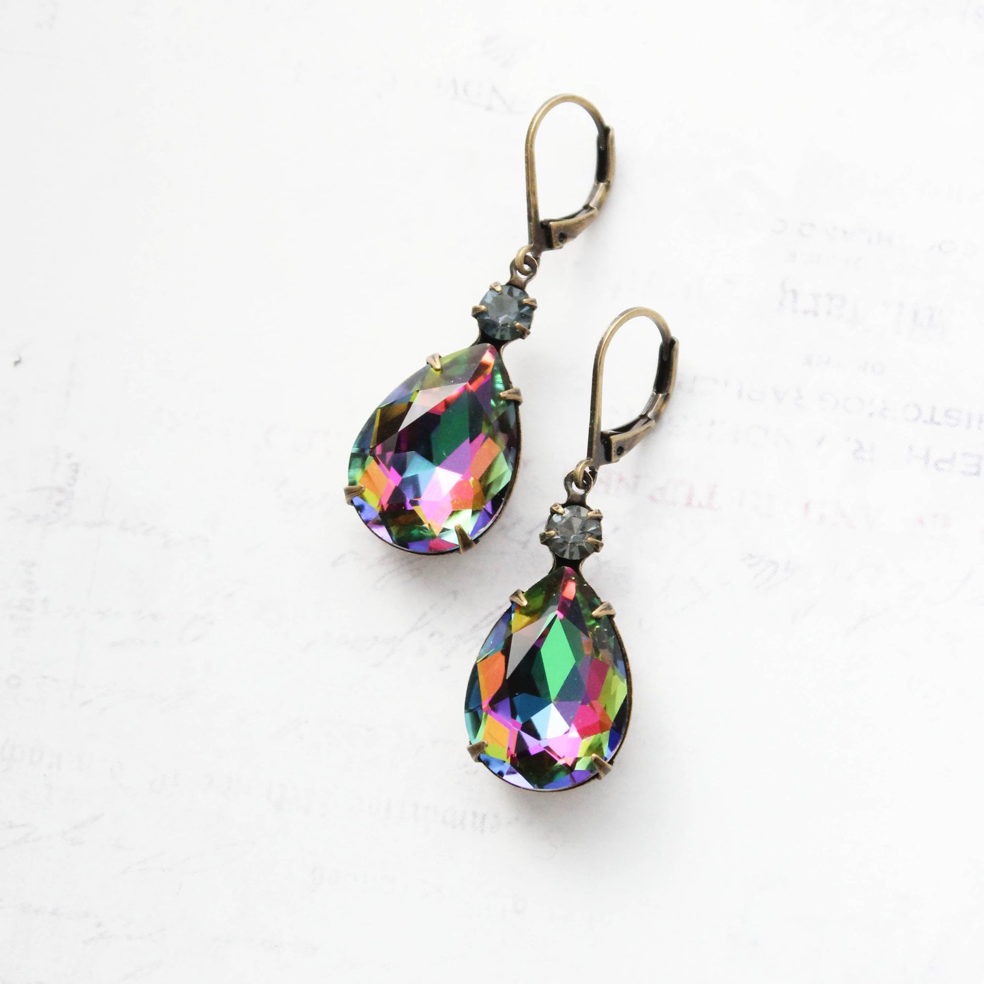 A pair of teardrop-shaped drop earrings with iridescent rainbow faceted crystals and antique bronze leverback hooks, displayed on a light textured white background.