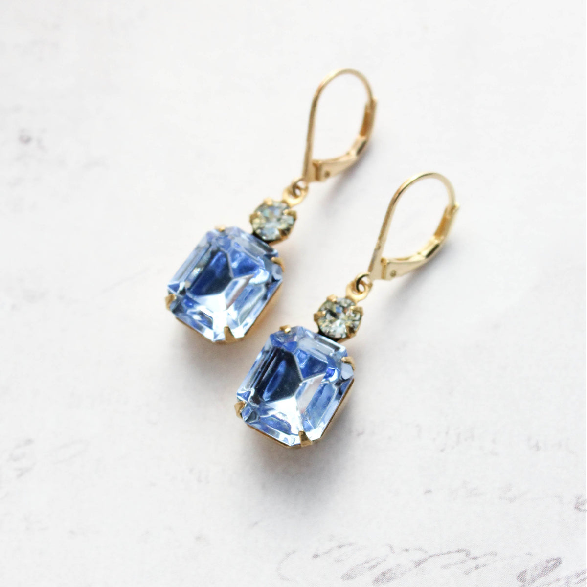 A pair of gold leverback drop earrings featuring light blue rectangular faceted gemstones, accented by small round clear stones above, set against a light textured background.