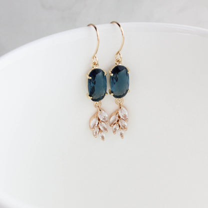A pair of gold drop earrings with dark blue oval stones and dangling leaf charms, resting inside a white ceramic cup.