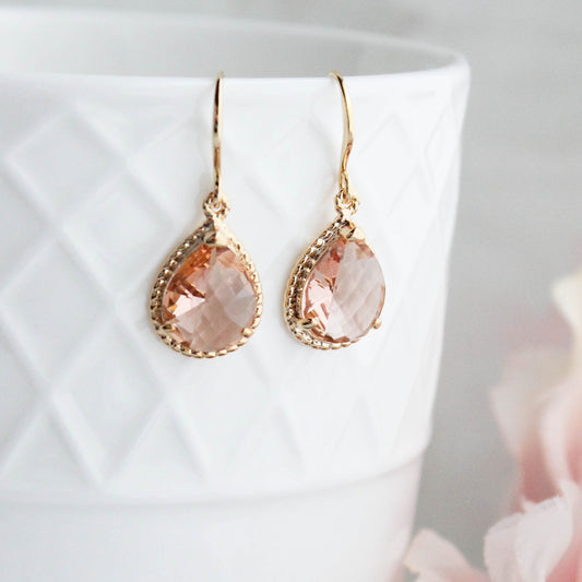 A pair of gold drop earrings with teardrop-shaped peach-pink faceted gemstones, resting on a white textured ceramic cup with soft pink flowers in the foreground.