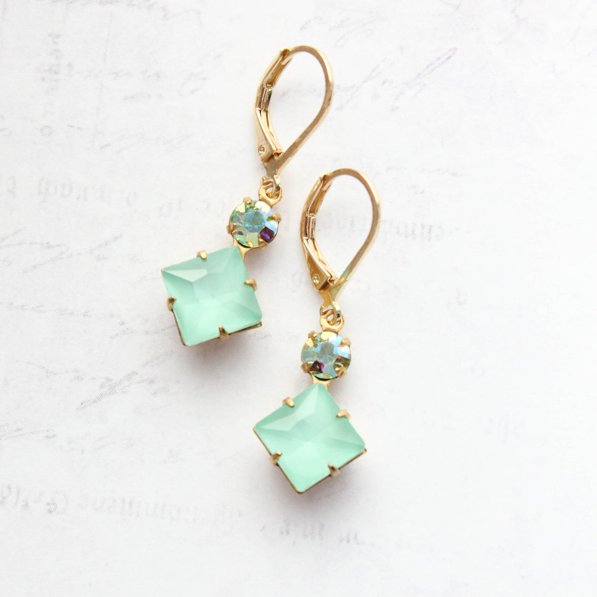 A pair of gold leverback drop earrings featuring square mint green gemstones and round iridescent blue-green crystals, displayed on a light textured background.