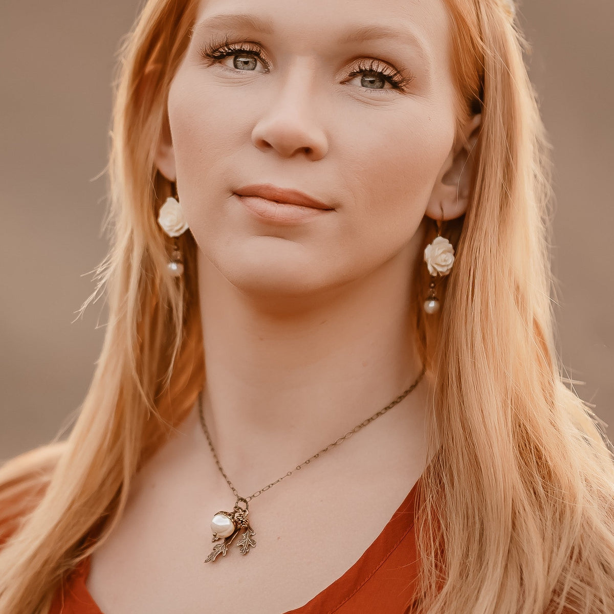 A close-up portrait of a young woman with long blonde hair, wearing a delicate gold necklace with a pearl and leaf pendant, matching drop earrings, and a rust-orange top, set against a warm, blurred background.
