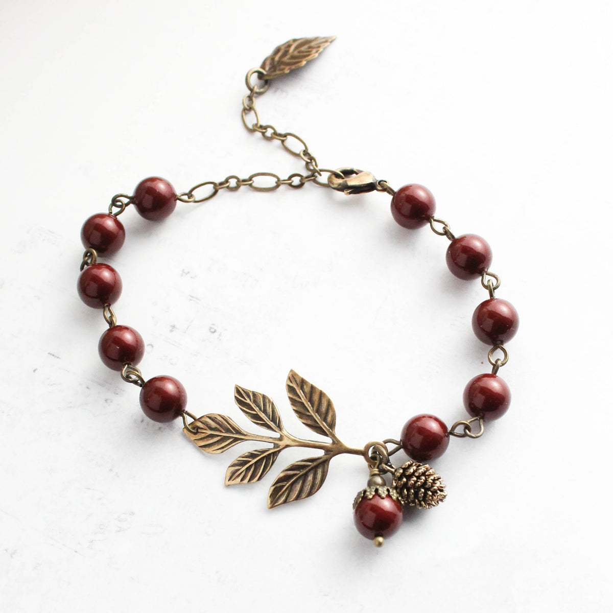 A beaded bracelet with dark red round beads, an antique bronze leaf branch centerpiece, and a small pinecone charm, displayed on a light textured background.