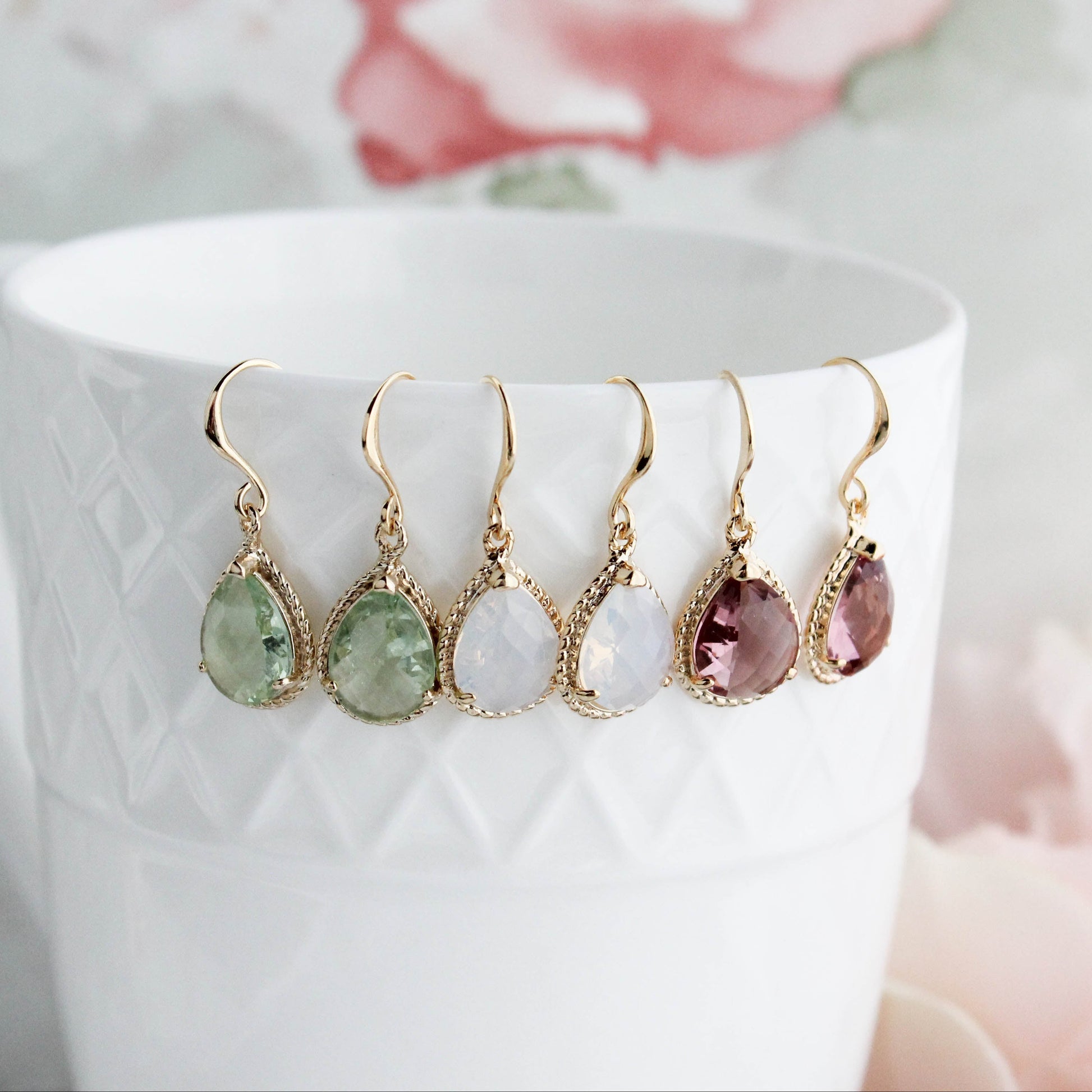 Six gold-tone drop earrings with teardrop-shaped gemstones in green, white, and dusty rose, hanging from the rim of a white textured ceramic mug against a soft pink floral background.