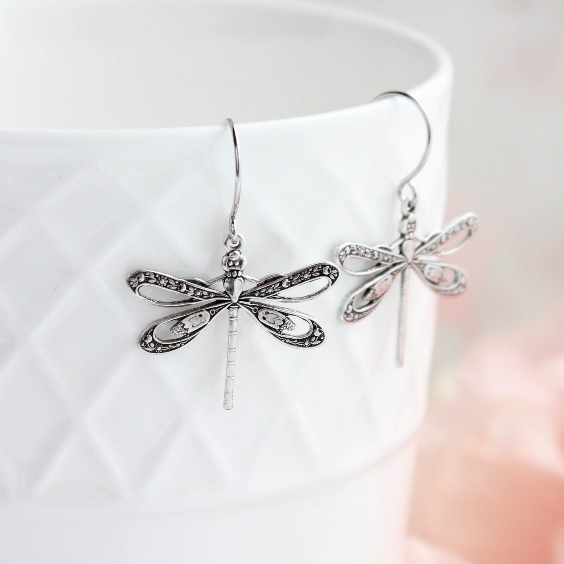 A pair of silver dragonfly drop earrings with delicate filigree detailing, hanging from hooks and displayed against a white textured ceramic cup with a soft pink blurred background.