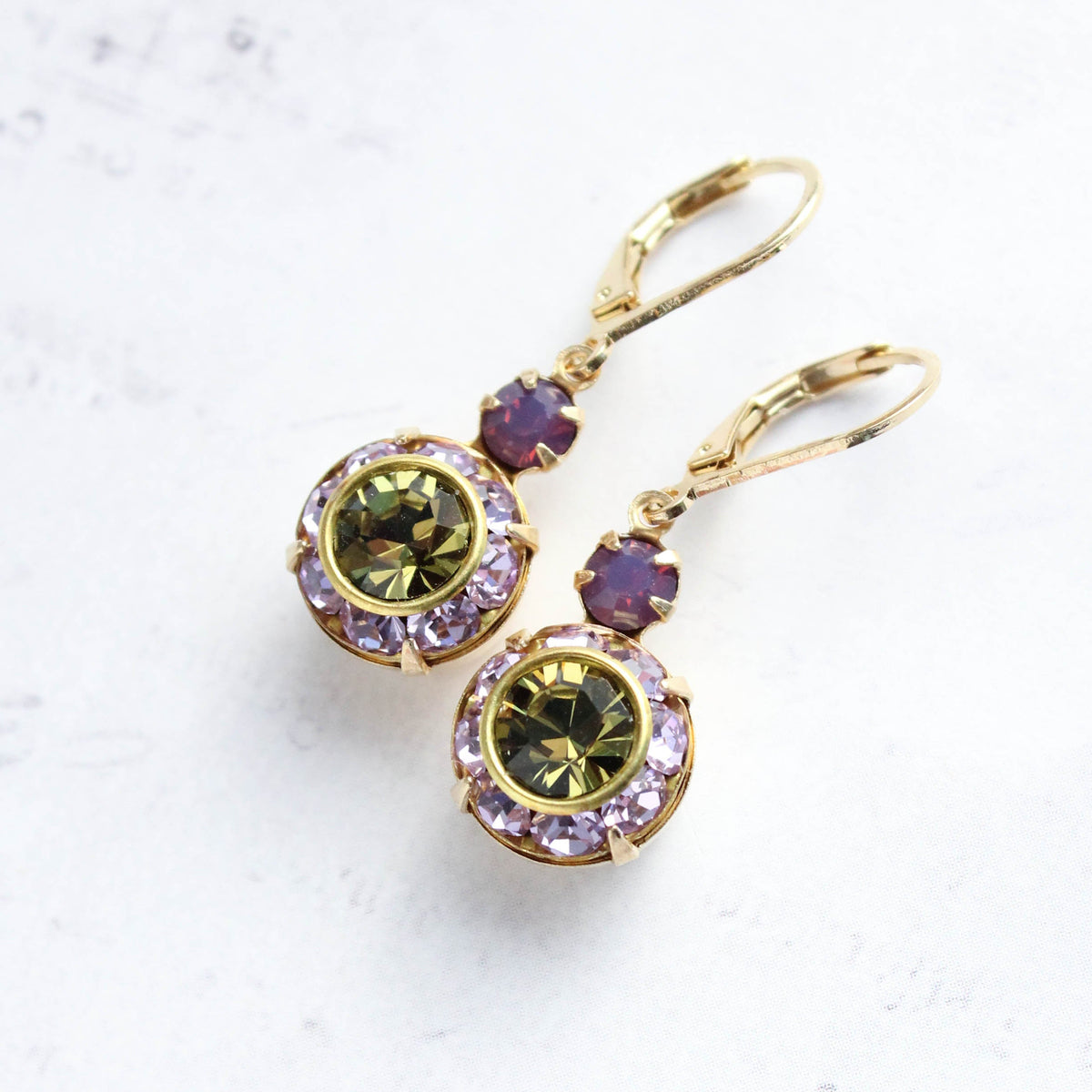 A pair of gold leverback earrings featuring round bezel-set olive green crystals surrounded by small lavender crystals, with smaller purple accent stones above, displayed on a light textured background.