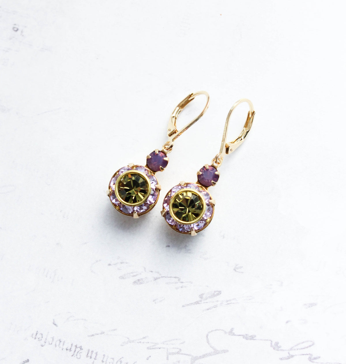 A pair of gold leverback drop earrings featuring oval green gemstones surrounded by small lavender crystal accents, displayed on a light textured background.