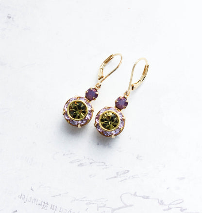 A pair of gold leverback drop earrings featuring oval green gemstones surrounded by small lavender crystal accents, displayed on a light textured background.