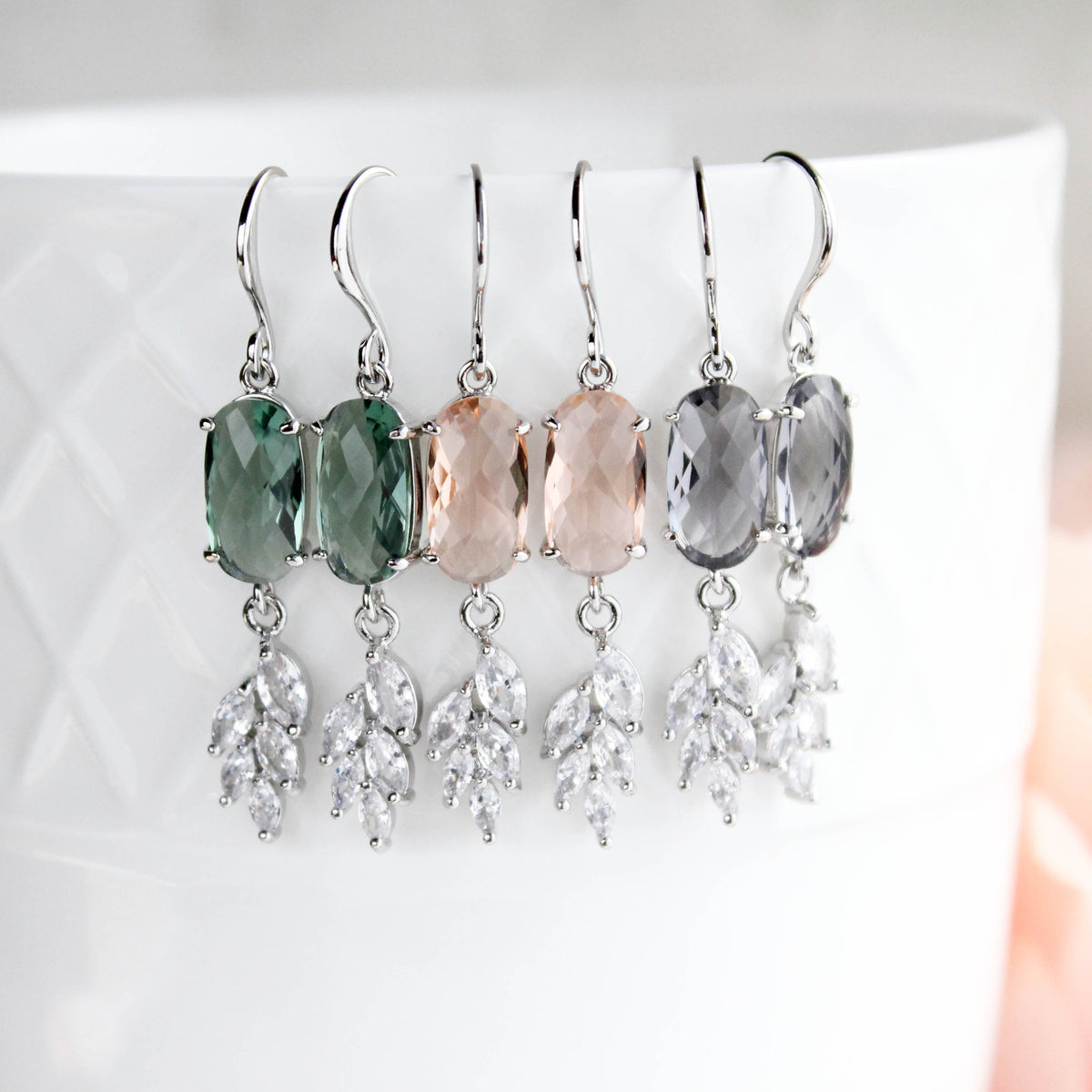 three pairs of dangling earrings displayed on a white textured ceramic cup. Each pair features a faceted oval top stone in green, blush pink, or smoky gray, suspended from silver hooks with leaf-shaped clear crystal drops.