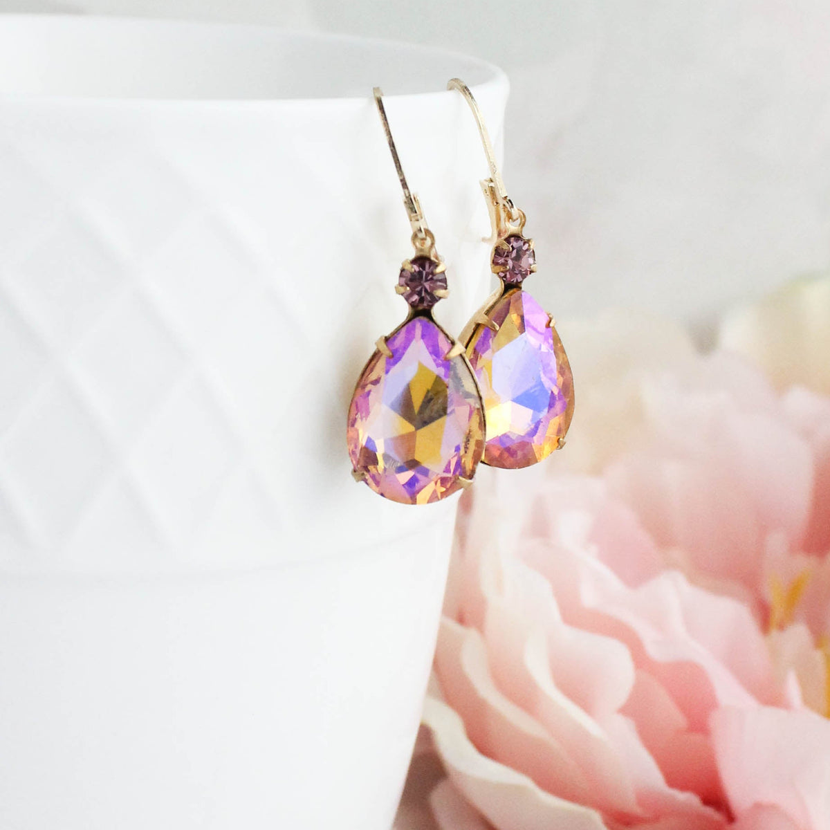 A pair of gold-tone teardrop dangle earrings with iridescent purple and pink faceted crystals, hanging from the rim of a white textured ceramic cup with soft pink flowers in the background.