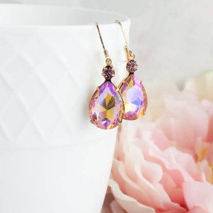 A pair of gold-tone teardrop dangle earrings with iridescent purple and pink faceted crystals, hanging from the rim of a white textured ceramic cup with soft pink flowers in the background.