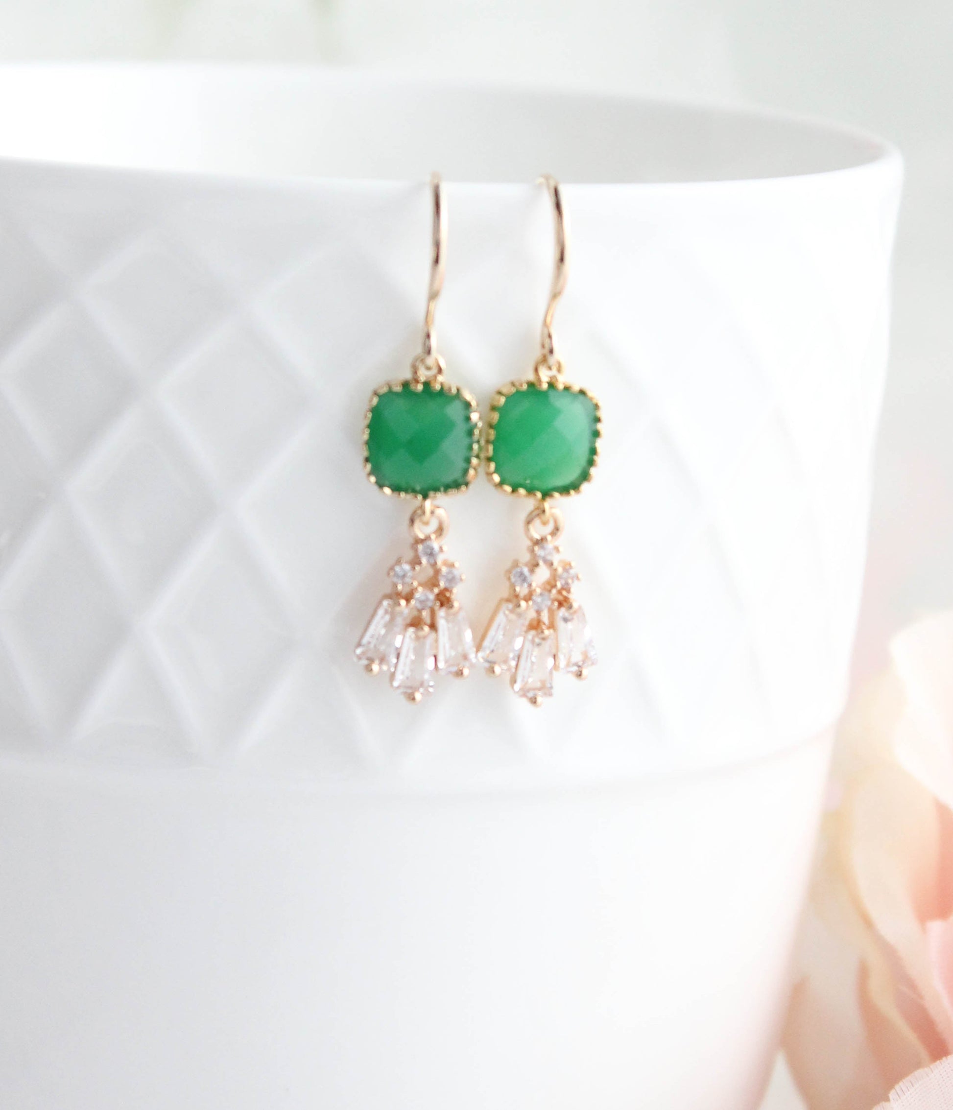 A pair of gold-tone dangle earrings with green rectangular stone tops and clear teardrop-shaped crystals, displayed on a white textured ceramic cup with a soft pink flower in the background.