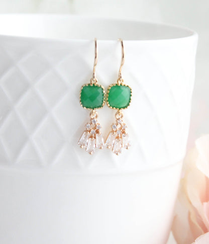 A pair of gold-tone dangle earrings with green rectangular stone tops and clear teardrop-shaped crystals, displayed on a white textured ceramic cup with a soft pink flower in the background.