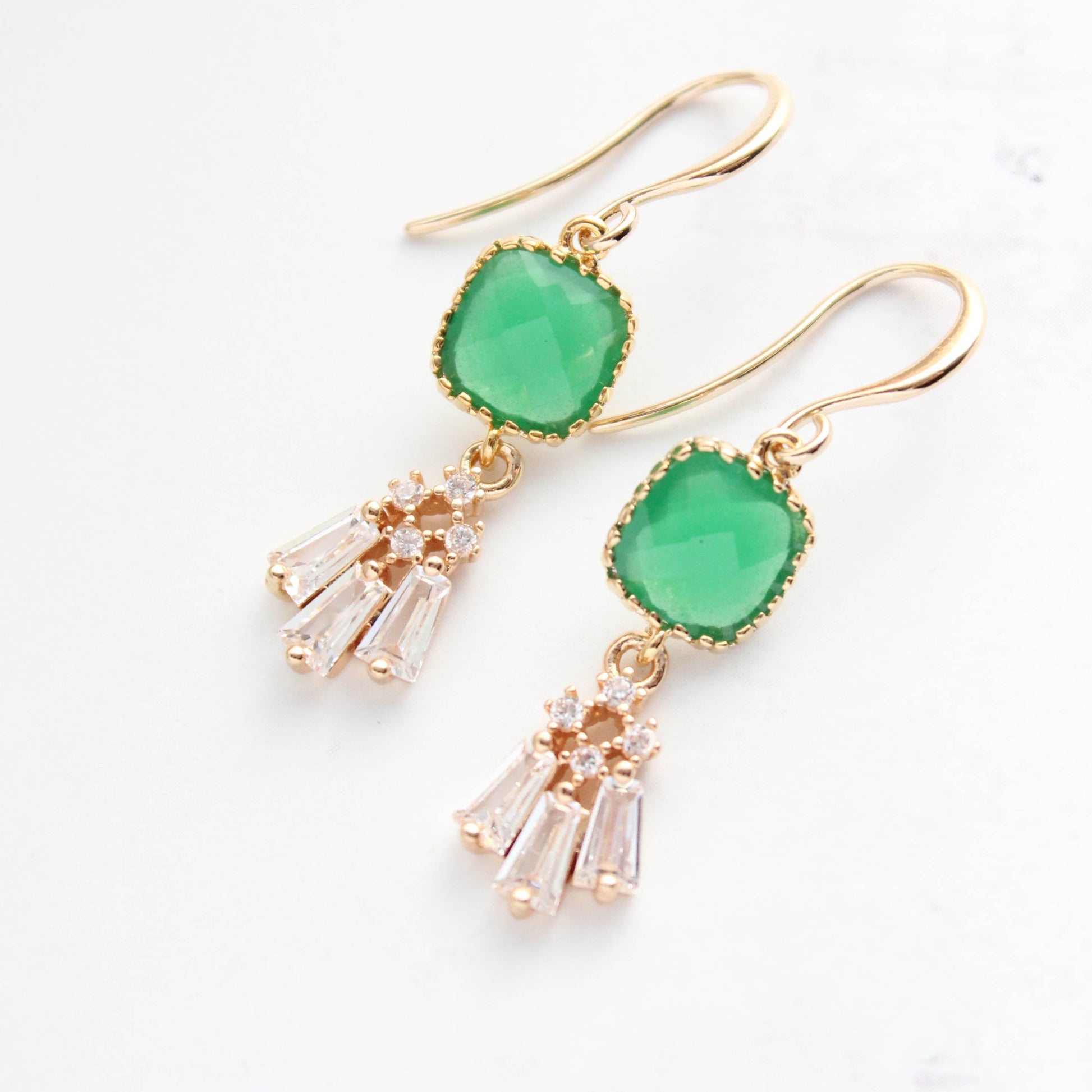 A pair of gold dangle earrings featuring a green cushion-cut stone and a dangling cluster of clear crystal-like drops, shown on a light off-white background.