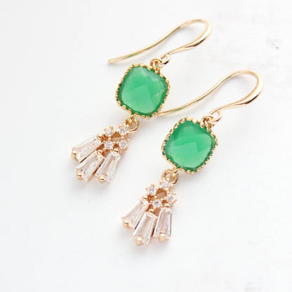 A pair of gold dangle earrings featuring a green cushion-cut stone and a dangling cluster of clear crystal-like drops, shown on a light off-white background.