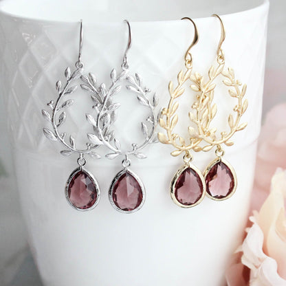 Two pairs of earrings - one in silver and one in gold - both are leaf wreath drop earrings with teardrop-shaped dusty rose plum glass jewels on the bottom, displayed on a white textured ceramic cup with pink flowers in the background.