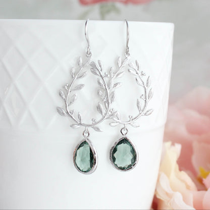 A pair of silver drop earrings with leafy wreath designs and teardrop-shaped green gemstones, displayed on a white textured ceramic cup with soft pink flowers in the background.