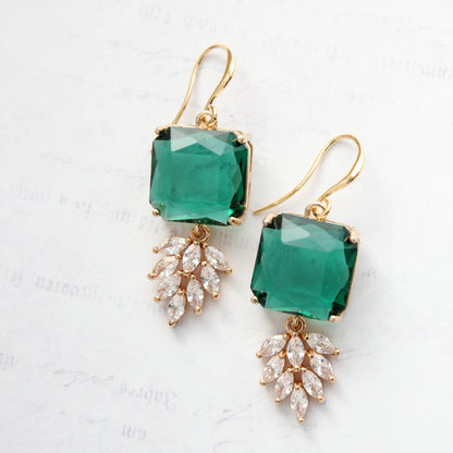 A pair of gold drop earrings featuring square green gemstones and leaf-shaped clear crystal clusters, displayed on a light textured background.