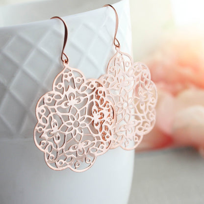 Rose gold laser-cut filigree dangle earrings with floral patterns, shown hanging from a white textured ceramic cup with a soft-focus pink background.