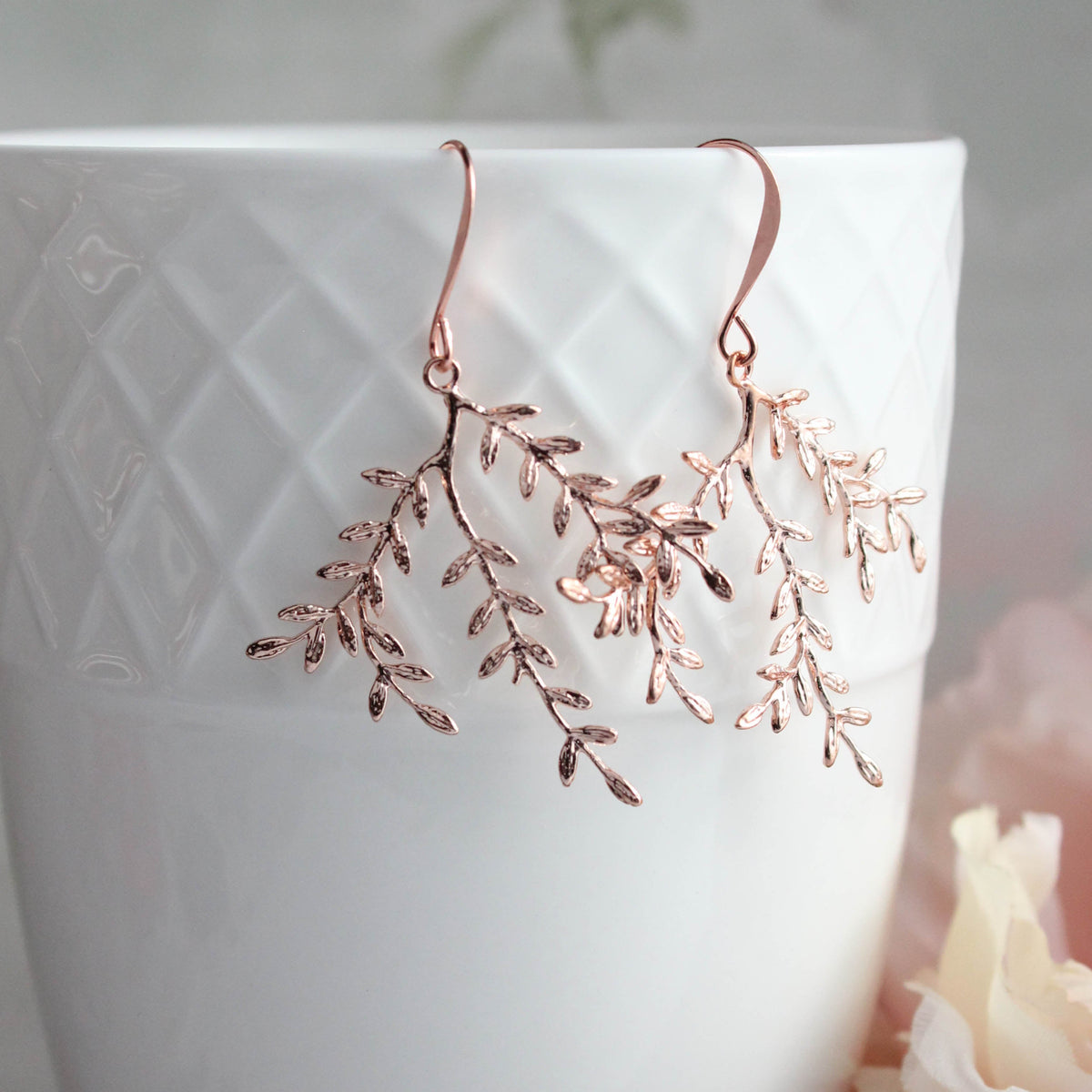 Rose gold branch drop earrings hanging from a white textured ceramic cup with soft pink flowers in the background.