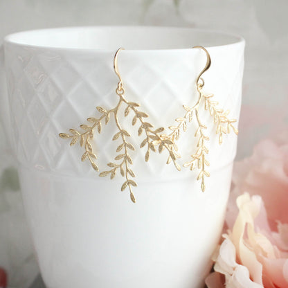 A pair of gold leafy branch dangle earrings hanging from a white textured ceramic cup, with soft pink flowers blurred in the background.