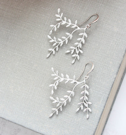 A pair of silver-toned dangle earrings with delicate leafy branch designs, displayed on a textured light gray fabric surface.