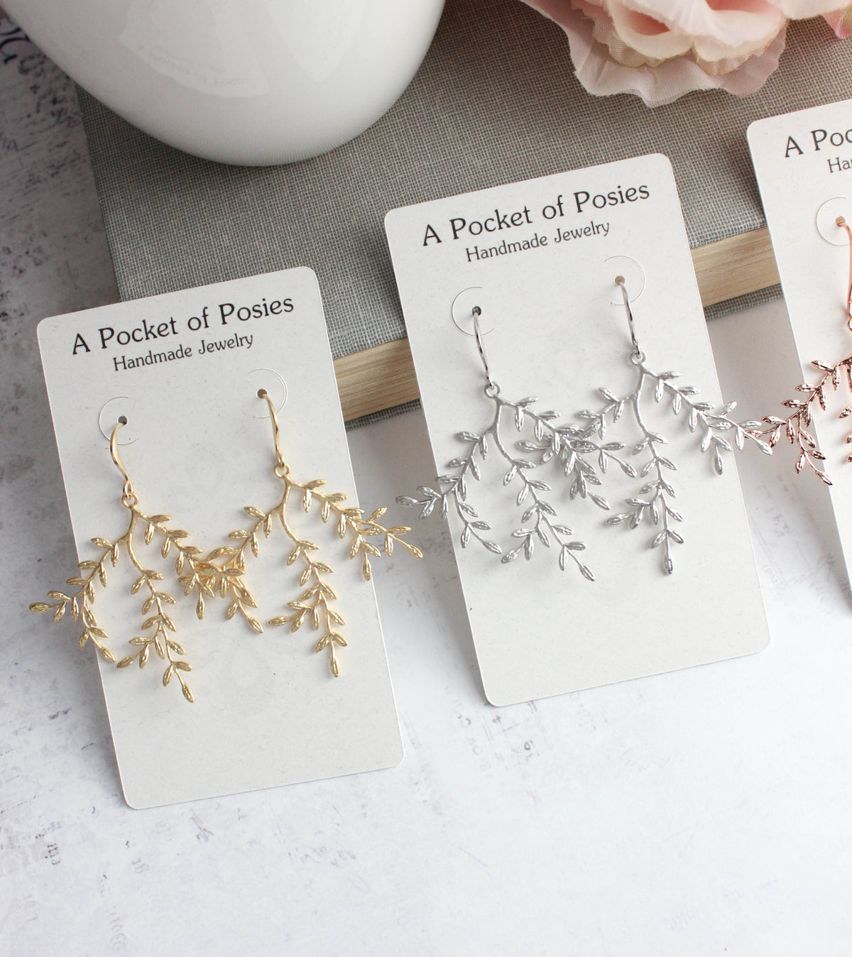 Three pairs of delicate branch-shaped dangle earrings displayed on white cards. The left pair is gold, the middle pair is silver, and a partial pink pair is visible on the right.