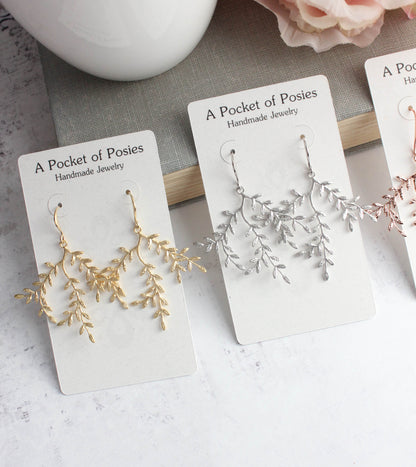 Three pairs of delicate branch-shaped dangle earrings displayed on white cards. The left pair is gold, the middle pair is silver, and a partial pink pair is visible on the right.