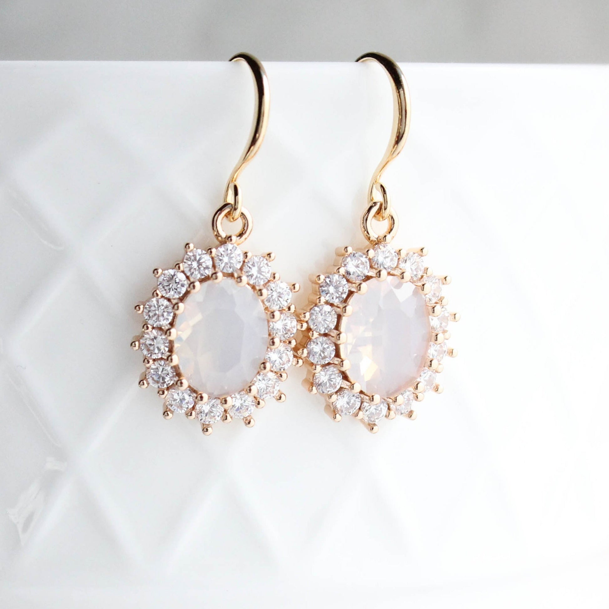 Gold earrings with an oval opal glass jewel in the center and small crystal glass jewels framing the center stone.  These earrings are handing on a white background.