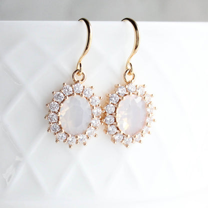 Gold earrings with an oval opal glass jewel in the center and small crystal glass jewels framing the center stone.  These earrings are handing on a white background.