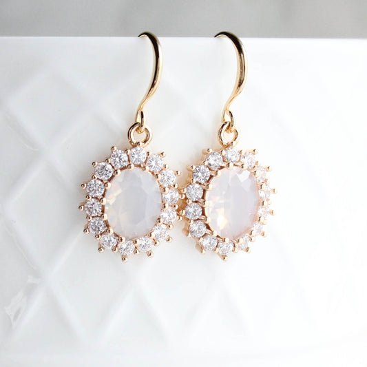 Gold earrings with an oval opal glass jewel in the center and small crystal glass jewels framing the center stone.  These earrings are handing on a white background.