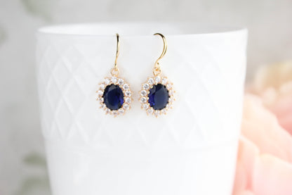 A pair of gold-tone dangle earrings with oval dark blue center stones surrounded by small clear stones, displayed against a white textured cup with a soft pink floral background.