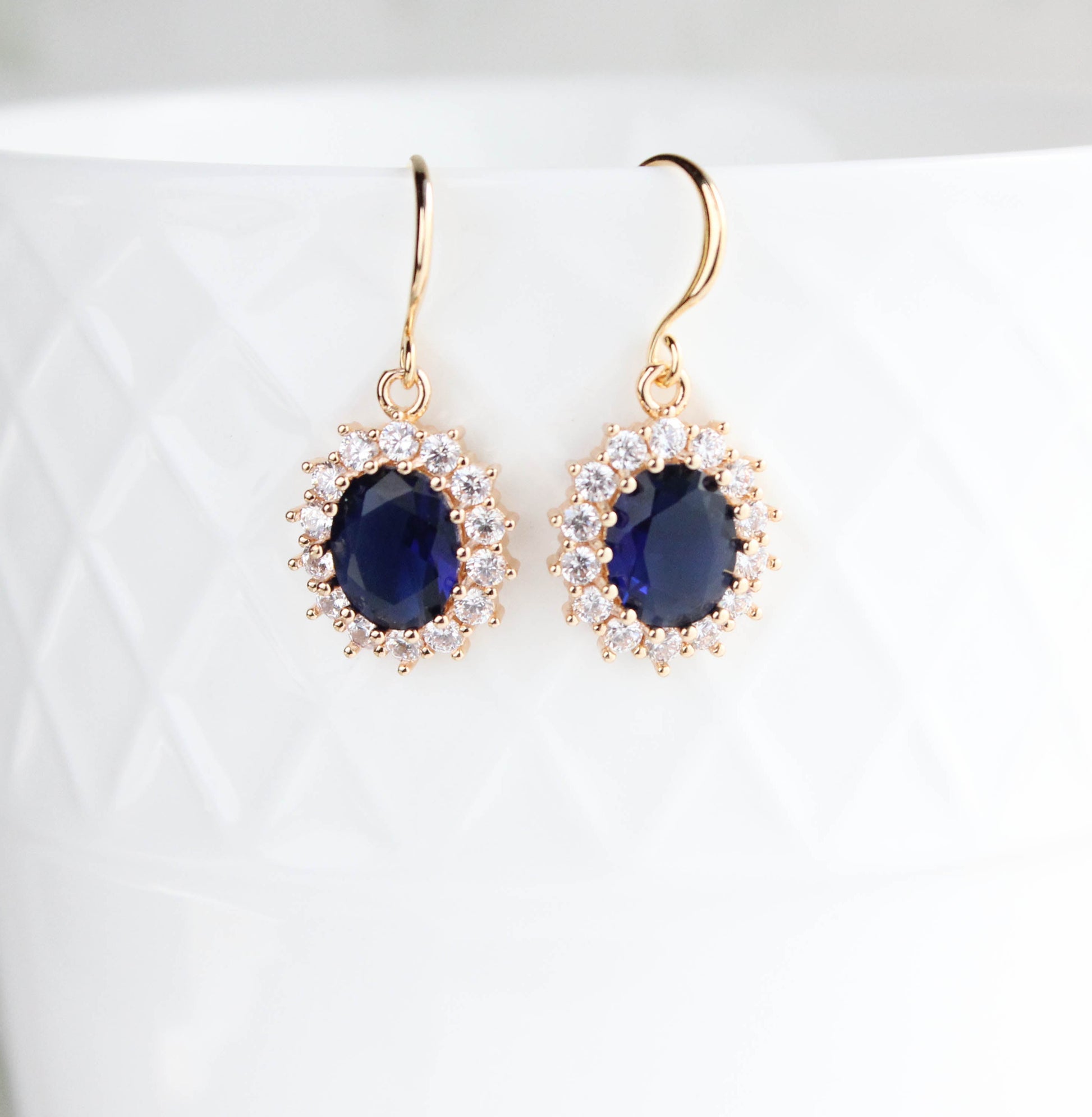 A pair of gold drop earrings with oval dark blue gemstones surrounded by small clear stones, displayed against a white textured ceramic background.