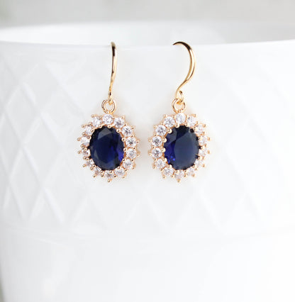 A pair of gold drop earrings with oval dark blue gemstones surrounded by small clear stones, displayed against a white textured ceramic background.